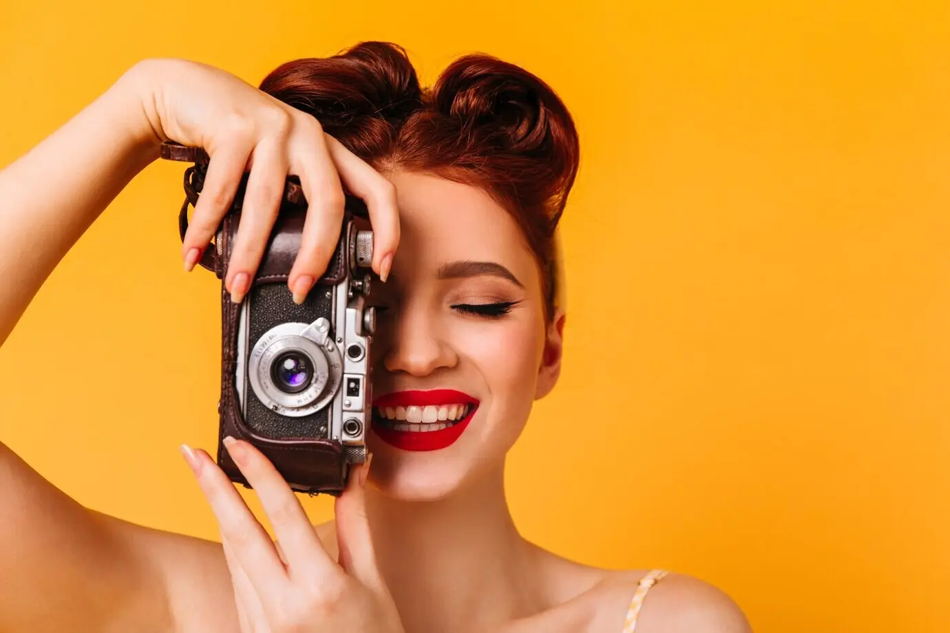 Joyful pin-up girl taking photos. Studio portrait of a woman with a camera, isolated against a yellow background.